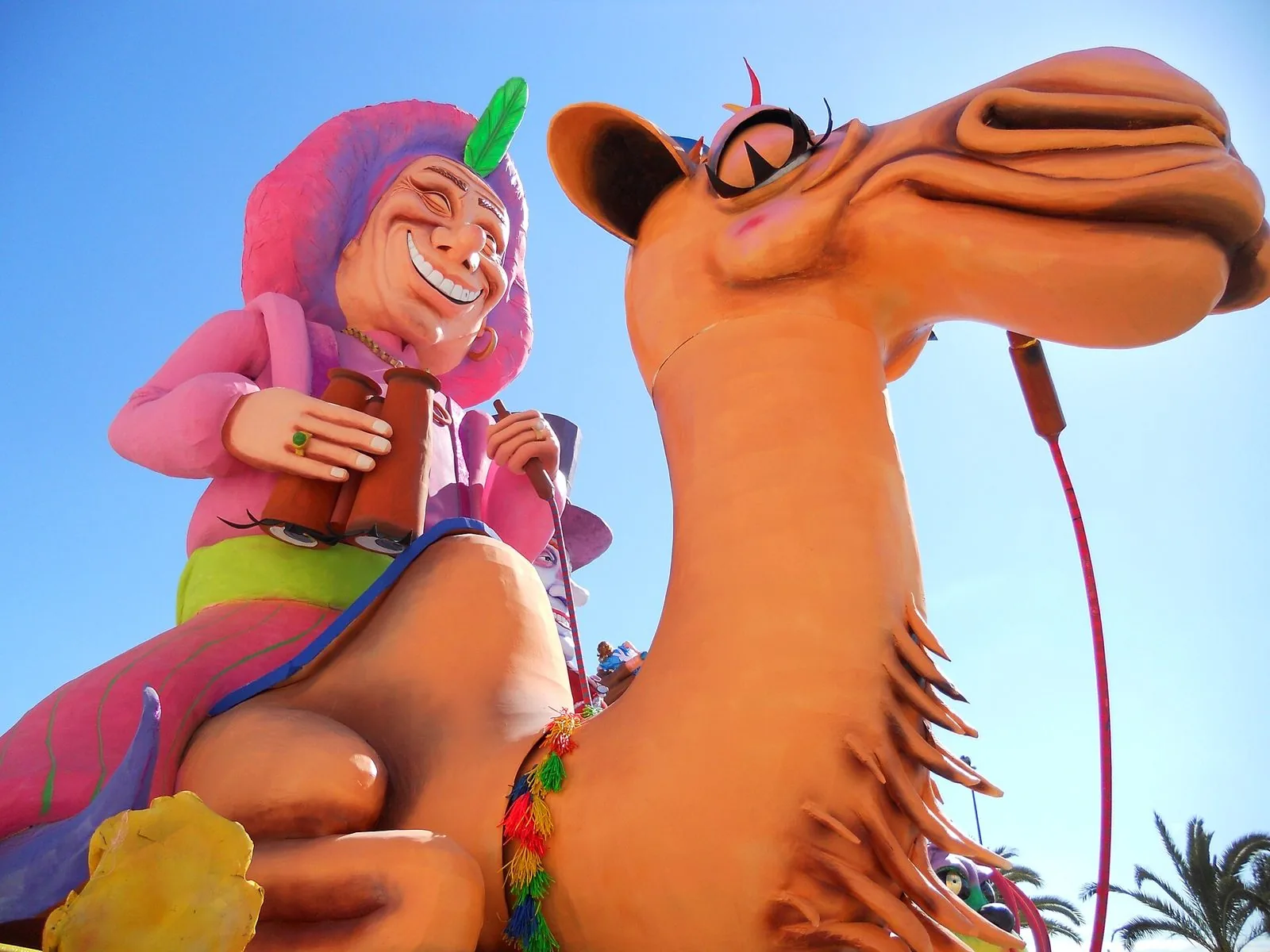 Carro allegorico di cartapesta del Carnevale di Manfredonia: una signora sorridente con binocolo in sella a un grande cammello, cielo azzurro e palme