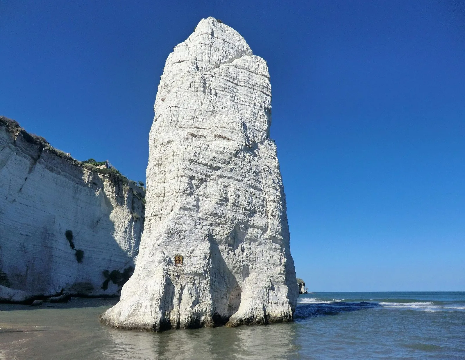 Il monolite bianco del Pizzomunno sulla spiaggia di Vieste, accanto alla falesia calcarea, con il mare calmo e il cielo azzurro