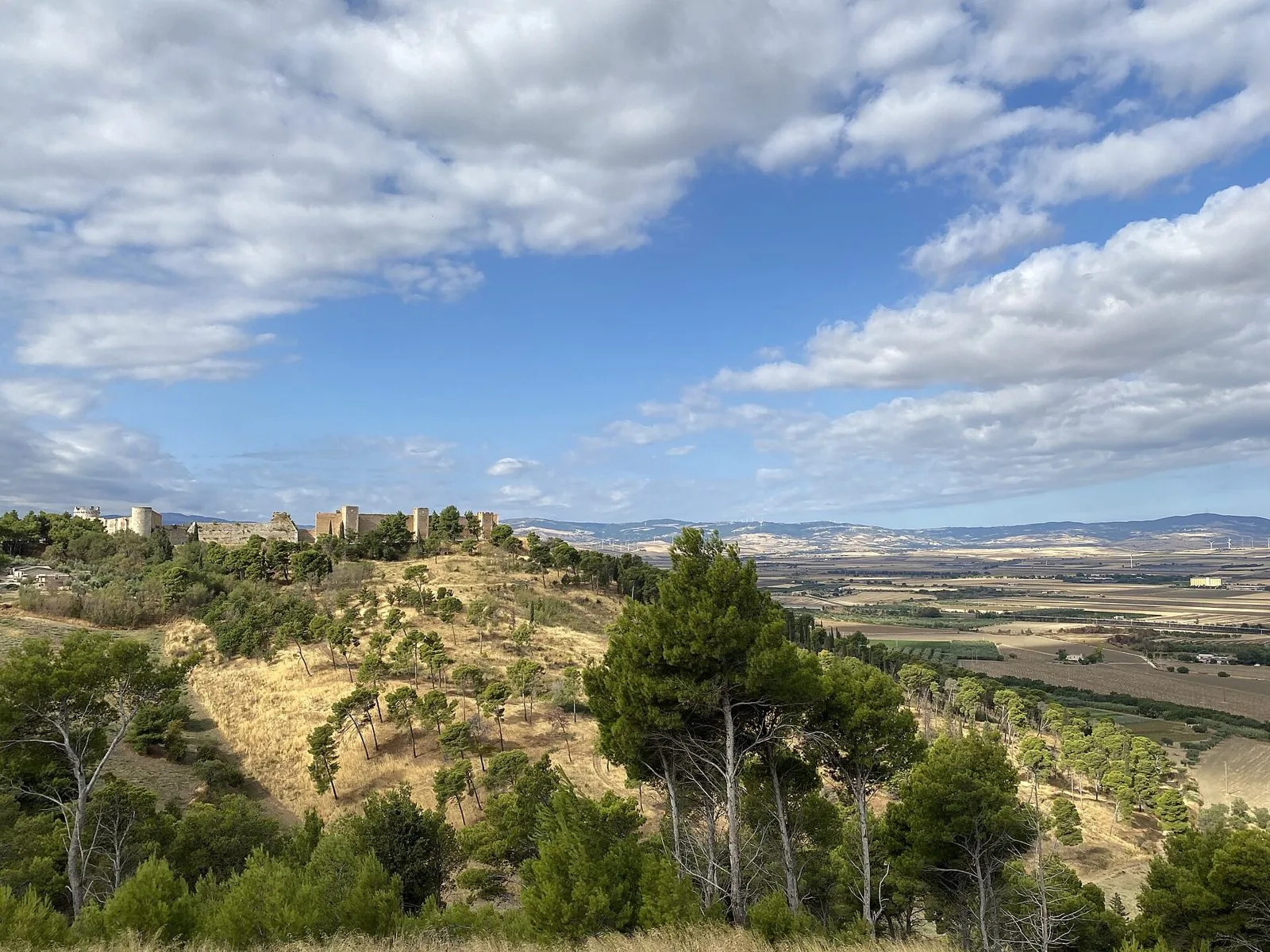 Le mura e le torri della fortezza di Lucera in cima a una collina con pini, sullo sfondo la pianura del Tavoliere e i Monti Dauni